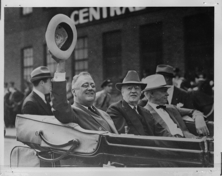 Black and white pohto of President Franklin Roosevelt in a convertible, riding in a parade, waving his hat to bystanders.
