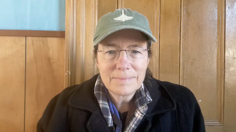 Caucasian female with dark hazel eyes, light brown hair tied in a loose ponytail, in her mid fifties, wearing frameless glasses, and donning faded aqua cap with an winged emblem, and wearing a heavy fleece jacket over a plaid button-down.