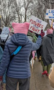 The backside of a person in a pink knitted hat with pointy corners on the left and right side, holding a sign displaying: Our futures our fight.