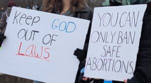 Protest sign displaying: Keep God out of laws. And another sign displaying: You can only ban safe abortions.