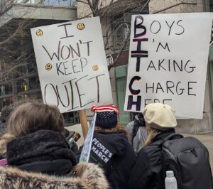Protestors holding signs. One displays: I won't keep quiet. Another displays: Boys I'm taking charge here.