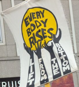Protest sign displaying: Four arms holding a ball. Each arm has one of the following words, Lift, From, The, and bottom. The ball displays: Every body rises.