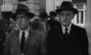 Black and white vintage image of two men standing shoulder to shoulder in fedora hats and suits with ties.