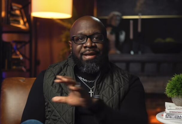 African American with dark rimmed glasses sitting in a dimly lit den in mid speaking.